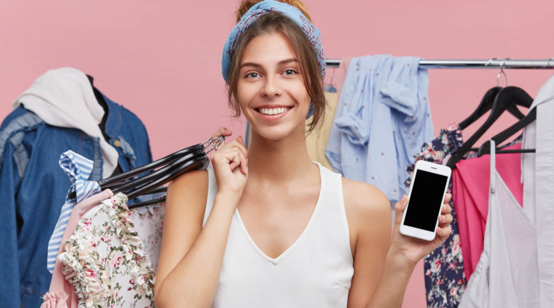 Retail, sale, consumerism and modern technology concept. Portrait of charming young female standing at rack with fashionable clothes, enjoying shopping at mall, paying with online app on cell phone