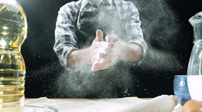 Professional male cook sprinkles dough with flour, preapares or bakes bread or pasta at kitchen table, has dirty uniform, isolated over black chalk background. Baking concept